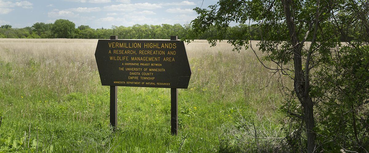 The Vermillion Highlands identification sign stands in front of an open, grassy area. There is a tree to the right of it and in the distance.